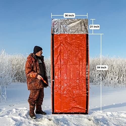 Person standing next to an orange and silver emergency survival tent in snowy landscape with dimensions marked.